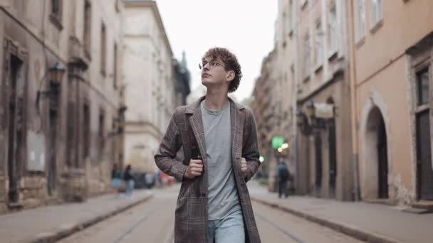 Joven millennial en gafas con abrigo a cuadros y pantalones vaqueros en auriculares con bolsa caminando, mirando a su alrededor en Old City Street. Estudiante, Concepto Turístico. Viajar . — Vídeo de stock