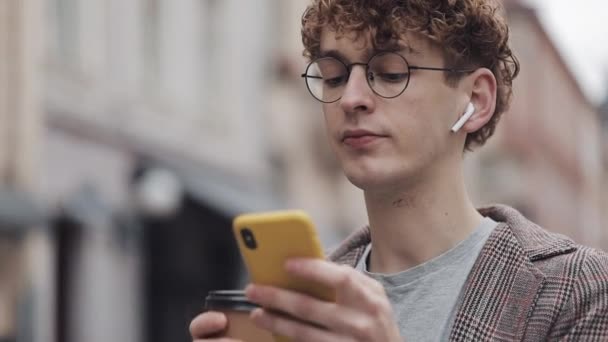 Close Up of Young Hipster Guy in Glasses and with Earphones Wearing Jacket Using His Smartphone Typing Message in Social Networks, Drinking Coffee Standing at City Street. — Stock video