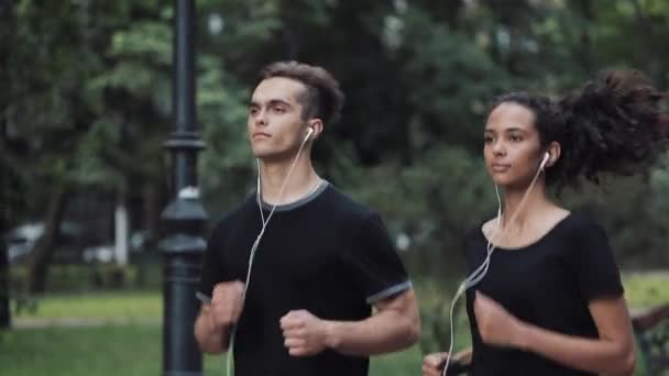 Красивый молодой человек и симпатичная мышиная девочка в наушниках выглядят расслабленными, бегая в парке . — стоковое видео
