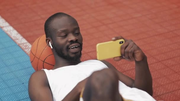 Close Up of Afro American Guy in Earphones Lying on with Ball under his Head, Watching Online Game on his Smartphone, Looking Extremily Happy and Rejoicing at Basketball Court. Sport Concept. — Stock Video