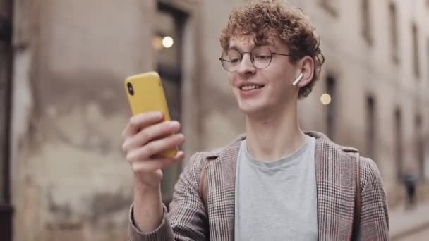 Close Up of Millennial em fones de ouvido e óculos Vídeo conversando com amigos, conversando, rindo via telefone móvel moderno, andando no fundo da Old City Street. Conceito de Comunicação . — Vídeo de Stock
