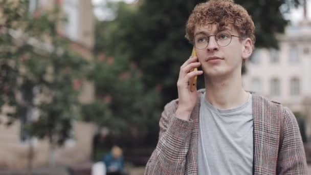 Крупный план молодого симпатичного парня в очках и пиджаке, разговаривающего по мобильному телефону на фоне Сити-стрит. Ученик . — стоковое видео