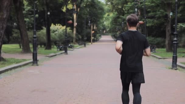 Молода людина з Кавказу в навушниках, яка носить костюм для бігу в парку Погляд на концепцію здорового способу життя. — стокове відео