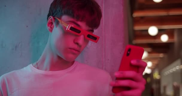 Mladý frajer ve stylových moderních brýlích stojí opřený o zeď a používá svůj chytrý telefon, dívá se na obrazovku na Futuristic Neon Club Lights. Zavřít snímek. — Stock video