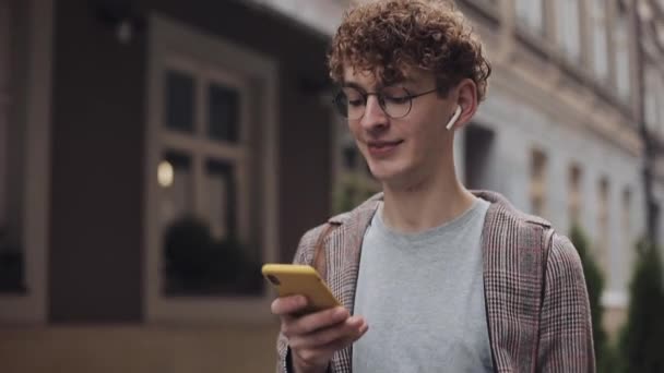 Close Up del giovane ragazzo hipster sorridente in occhiali e auricolari che indossa giacca a quadri, comunica con gli amici nei social network, Utilizzando il suo moderno telefono cellulare, Camminando lungo Old City Street . — Video Stock