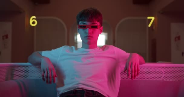 Cool Young Trendy Guy w białej koszulce w stylowych okularach siedzi zrelaksowany na nowoczesnej kanapie patrząc prosto do kamery z futurystycznymi Neon Lighting Numbers w tle. — Wideo stockowe