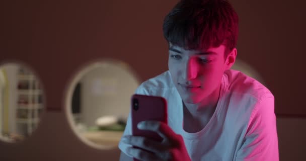 Κομψό Millenial Boy σε λευκό T-Shirt Χρησιμοποιώντας Smartphone του, Κοιτάζοντας την οθόνη και χαμογελώντας, ενώ στέκεται στο εσωτερικό Neon Φωτισμός και φουτουριστικό φόντο του δωματίου. — Αρχείο Βίντεο