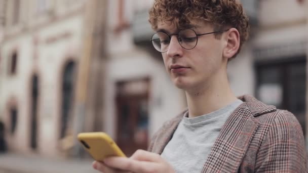 Fermer Portrait d'un jeune étudiant portant des lunettes à l'aide de son téléphone intelligent moderne, faisant défiler l'écran assis sur le fond de la rue Old City. Étudiant, Concept touristique . — Video