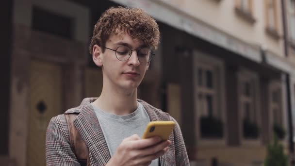 Πορτρέτο της Calm Young Hipster Man με κόκκινα σγουρά μαλλιά σε γυαλιά φορώντας σακάκι Χρησιμοποιώντας το κινητό του τηλέφωνο για να πληκτρολογήσετε το μήνυμα, ενώ στέκεται στο Old City Street Communication Concept. — Αρχείο Βίντεο