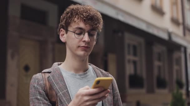 Portrét Klidný mladý hipster muž s červenými kudrnatými vlasy v brýlích nosit bundu pomocí svého mobilního telefonu psát zprávu, zatímco stojí na Old City Street Communication Concept. — Stock video