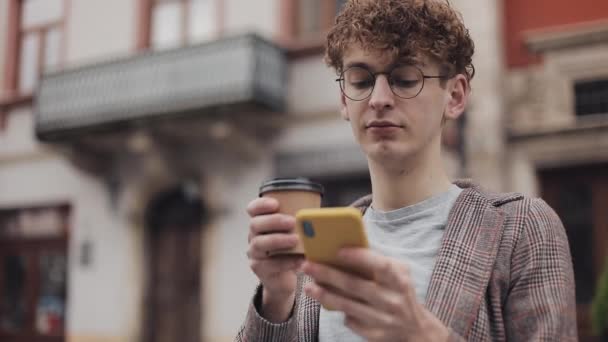 ジャケットと眼鏡を身に着けている若いヒップスター男は、コーヒーを飲みながら、シティストリートのベンチに座っている間、彼の現代的な携帯電話の画面をスクロールします。学生概念.Bottom view. — ストック動画