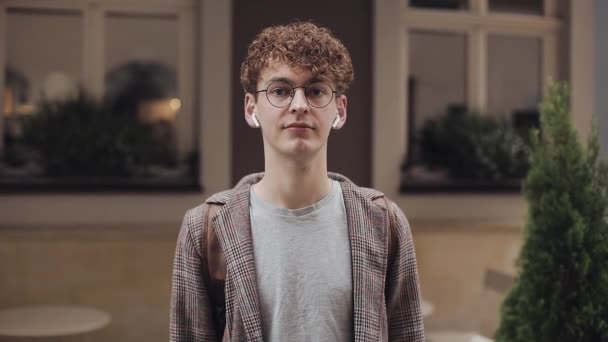 Portrait de jeune homme avec des cheveux bouclés rouges dans les lunettes portant une veste à carreaux et avec des écouteurs, regardant vers la caméra, debout fond de la vieille ville. Étudiant, Concept touristique . — Video