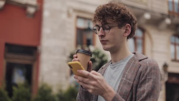 Rechte Seitenansicht eines jungen, gut aussehenden Studenten mit Brille, der karierte Jacken trägt und auf seinen Smartphone-Bildschirm schaut, Kaffee trinkt und vor Altstadthintergrund spaziert. Touristen. — Stockvideo