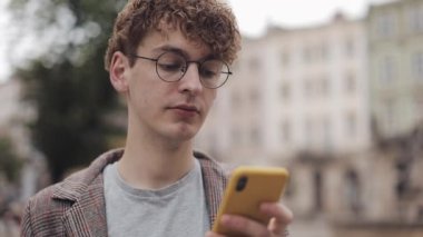 Close Up Portrait of Serious Millennial in Glasses Using his Modern Mobile phone App,Looking at Screen, Drinking Coffee, at Blured City Background.