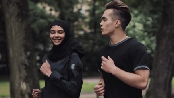 Jeune homme caucasien et femmes mousseline portant le hijab regardant excité et détendu souriant et bavardant courir dans le parc vue de côté . — Video