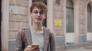 Kızıl Kıvırcık Saçlı Gözlüklü Genç Adam 'ın portresi Gözlüklü Ceket Giyiyor ve Smaertphone ile, Kameraya Bakıyor, Eski Şehir Arkaplanı Duruyor. Öğrenci, Turist Konsepti.