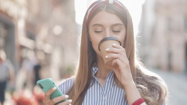 Ritratto di bella giovane ragazza caucasica con capelli castani e fascia che indossa camicia a righe utilizzando il suo smartphone e sorridente bere caffè in piedi sullo sfondo della città . — Video Stock