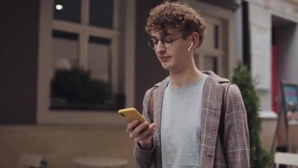 Giovane ragazzo hipster in occhiali con gli auricolari indossa giacca a quadri avendo zaino. Chiacchierare con gli amici, Utilizzando il suo smartphone. Camminare sullo sfondo della città vecchia. Studente, concetto turistico. — Video Stock