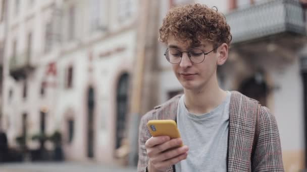 Joven estudiante nerd en gafas usando su dispositivo de teléfono móvil moderno para chatear con amigos, escribiendo en la pantalla de pie al aire libre. Comunicación, Estudiante, Concepto Turístico . — Vídeo de stock