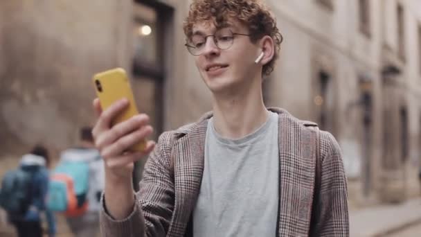 Millennial in Kulaklıklar ve Gözlük Giyen Ceket Giyen Arkadaşlarla Gülüşen Sohbet Modern Cep Telefonu ile Merhaba, Eski Şehir Sokak Arkaplanında Yürüyüş. İletişim Kavramı. — Stok video