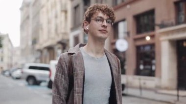Gözlüklü Genç Millennial, Kulaklıklı ve İşaretli Ceket ve Kot Giyiyor, Çanta Yürüyor, Eski Şehir Caddesine Bakınıyor. Öğrenci, Turist Konsepti. Seyahat.