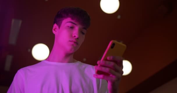 Der junge Millenium-Typ trägt ein weißes T-Shirt und sein Smartphone steht an futuristischen Neonlichtern. Kommunikations- und Technologiekonzept. untere Kameraeinstellung. — Stockvideo