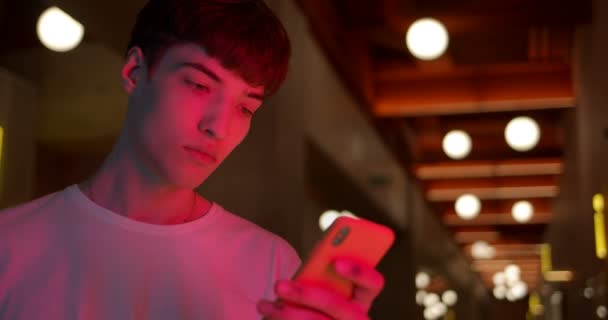 Νεαρός κομψός τύπος που φοράει λευκό μπλουζάκι Χρησιμοποιώντας το Smartphone του για να συνομιλεί στα κοινωνικά δίκτυα Στέκεται στο φουτουριστικό φόντο του Neon Club Lights. Κοντινό πλάνο. — Αρχείο Βίντεο