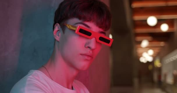 Retrato de un joven millenial con elegantes gafas de fiesta modernas de pie a un lado inclinado a la pared y mirando directamente a la cámara en Futuristic Neon Club Lights. Primer plano . — Vídeos de Stock