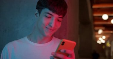Genç Stylish Millenial Boy 'un Fütürist Neon Kulüp Işıkları Arkaplanında Gülümseyen Smartphone' unu Kullanırken Yakın Çekimi. İletişim ve Teknoloji Konsepti.