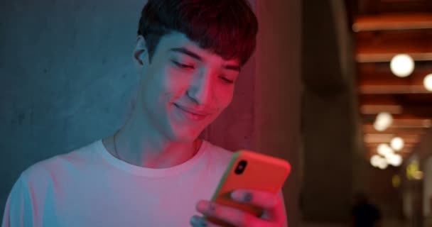 Close Up Shot of Young Elegante Millenial Boy Usando su Smartphone y Sonriendo de pie en Futuristic Neon Club Luces Fondo. Concepto de Comunicación y Tecnología . — Vídeos de Stock