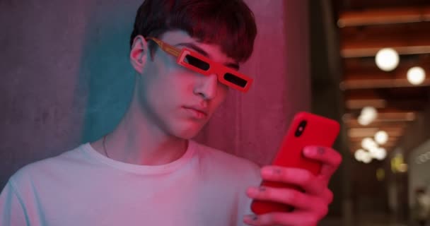 Young Millenial Guy in Stylish Modern Party Glasses Standing aside Leaned to Wall and Using his Smartphone, Looking to Screen at Futuristic Neon Club Lights. Close Up. — Stock Video