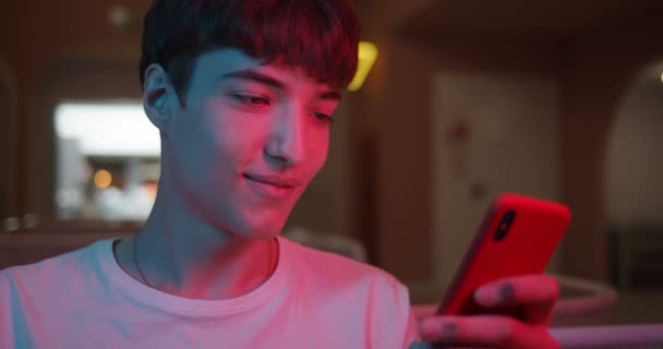 Close up colpo di elegante fresco Millenial Guy utilizzando il suo smartphone moderno e sorridente, chiacchierando nei social network mentre seduto al coperto con illuminazione al neon sullo sfondo . — Video Stock