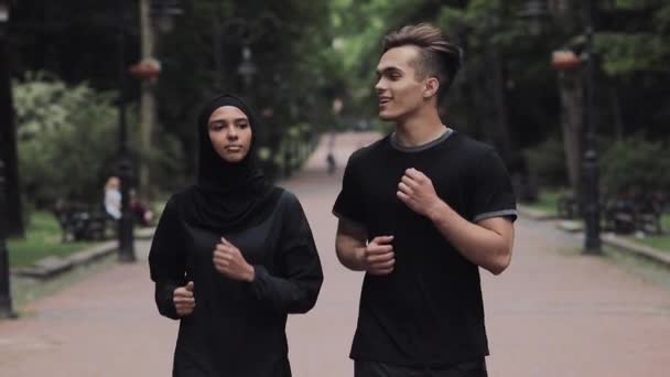Joven caucásico hombre y muselina mujeres usando hijab buscando excitado y relajado sonriendo y charlando corriendo en el parque . — Vídeo de stock