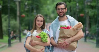 Yiyecek dolu kağıt torbaları taşırken kadın ve erkeğin gülümseyip kameraya baktığı görüntüler. Çekici kız ve gözlüklü sakallı erkek arkadaşı alışverişten sonra parkta yürüyorlar..