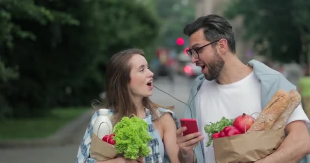 Boldog pár, akik jól érzik magukat, miközben zenét hallgatnak fejhallgatóban, és együtt énekelnek az utcán. A férfi és a felesége hazajönnek, miután bevásároltak és papírzacskóban vitték az ételt.. — Stock videók