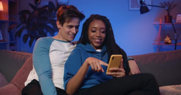 Junges Paar schaut aufs Smartphone und nutzt lustige Apps, während es die Zeit zusammen auf dem Sofa verbringt. Lächelnde Kerle und Mädchen entspannen sich und scrollen durch den Newsfeed der sozialen Medien. Freizeitkonzept. — Stockvideo