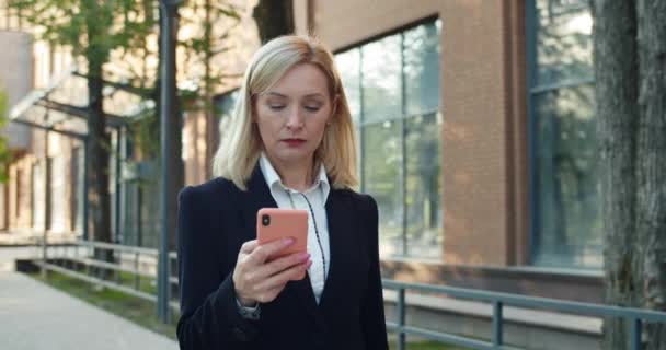 Serious woman in 30s scrolling and looking at smartphone screen while reading news. Beautiful businesswoman using her phone while walking at street near modern office building.