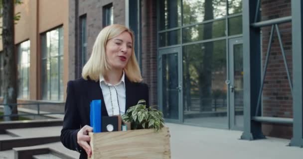 Atractiva empleada mujer en30s mirando alegre y sonriente, mientras que la obtención de un nuevo trabajo. Mujer feliz llevando y mirando sus cosas mientras camina y canta cerca de un edificio moderno . — Vídeos de Stock