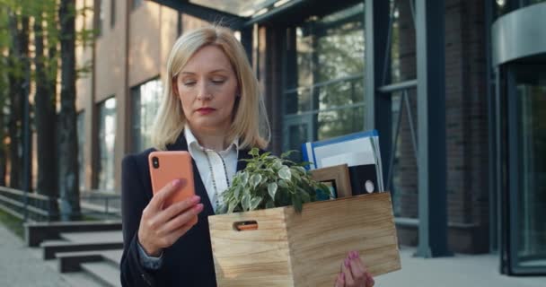 Mujer de negocios seria en ropa formal sosteniendo caja con sus cosas en la calle. Trabajadora de pie y desplazándose por la pantalla del teléfono inteligente mientras busca trabajo. Concepto de desempleo . — Vídeo de stock