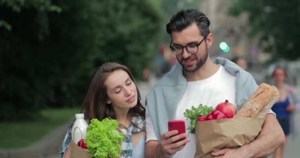 Hombres y mujeres alegres regresan a casa después de comprar y llevar comida en bolsas de papel. Pareja sonriente mirando la pantalla del teléfono inteligente y haciendo cara sorprendida mientras camina por la calle . — Vídeos de Stock