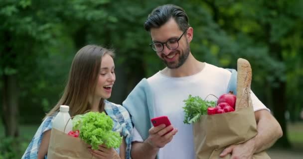 Cheerful couple looking at phone screen and laughing while carrying food in paper bags. Man showing his wife smartphone and talking while they smiling and walking through park. — Stock Video