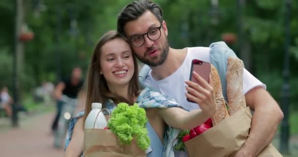 Pareja alegre sonriendo y posando para selfies con bolsas de papel llenas de comida. Hombre y mujer guapos en los años 30 haciendo fotos en la cámara frontal del teléfono inteligente mientras camina. Al aire libre . — Vídeos de Stock