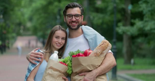 Портрет счастливой пары, смотрящей в камеру, держащей бумажные пакеты с едой и улыбающейся. Веселый мужчина в очках обнимает свою прекрасную жену, пока они позируют в парке. На открытом воздухе . — стоковое видео