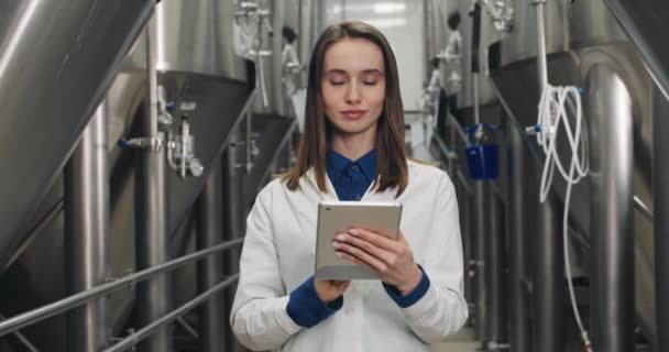 Retrato de la hermosa mujer ingeniero de procesos introduciendo datos en la tableta en la fábrica de cerveza. Mujer joven con bata de laboratorio blanca tocando y escribiendo la pantalla digital mientras está de pie y mirando a la cámara . — Vídeo de stock