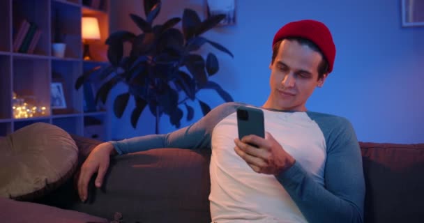 Sosyal ağlarda sohbet ederken akıllı telefonunu kullanan şık bir genç. Milenyum adamı günlük kıyafetlerle mesaj yazıyor ve kanepede otururken gülümsüyor. Ev arkaplanı. — Stok video