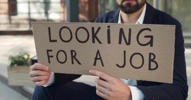 Widok upraw męskiego pracownika trzymającego baner kartonowy z szukając pracy pisania. Człowiek szukający pracy siedząc na ulicy. Pojęcie światowego kryzysu finansowego i bezrobocia. — Wideo stockowe