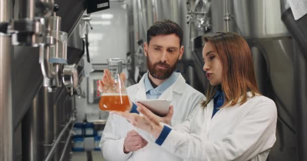 Pracownicy browaru męskiego i żeńskiego w fartuchach laboratoryjnych sprawdzają jakość produktu i rozmawiają. Mężczyzna z tabletką i kobieta trzymający szklaną kolbę z piwem patrząc na świeżo zrobiony napój. — Wideo stockowe