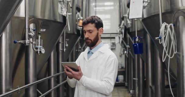 Ingeniero de procesos masculino guapo manteniendo registros en la tableta en la cervecería. Trabajador profesional tocando y tecleando la pantalla digital mientras camina y mira los portapapeles en el equipo . — Vídeo de stock