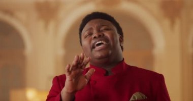 Kırmızı elbiseli genç Afro-Amerikalı adamın duygusal performansını yakından izleyin. Kilisede şarkı söyleyen erkek portresi. Müzik ve din kavramı.