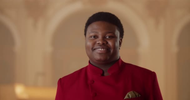 Portrait of cheerful young guy wearing red suit and looking to camera. Close up view of afro american guy in 30s turning head and smiling. Indoors.Blurred background. Concept of emotions. — Stock Video